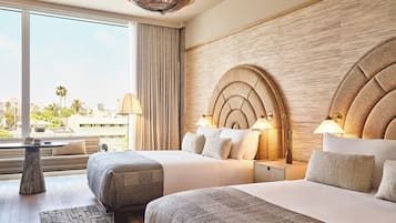 A hotel room with two beds, a large window with curtains, a wooden headboard, and a view of palm trees and buildings.