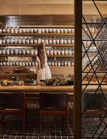Una mujer con un vestido blanco está de pie junto a un mostrador de madera con botes de especias. Hay dos sillas frente al mostrador y una ventana con un diseño geométrico.