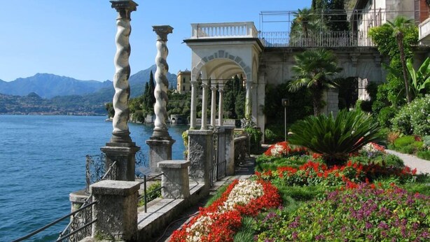 Un jardin au bord de l’eau, agrémenté de colonnes en pierre, d’un belvédère blanc et de fleurs aux couleurs vives.