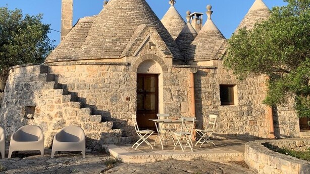 A stone building with multiple conical roofs and a central arched doorway. There is a small outdoor seating area with white chairs and a table. - PRICKLY PEAR - Cone Life (Cisternino)