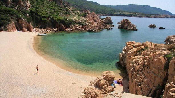 A beach with clear water, rocky cliffs, and a person standing on the sand.