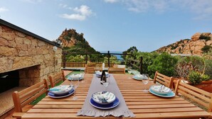 A wooden dining table set for four with plates, glasses, and a bottle, overlooking a rocky landscape and the sea.