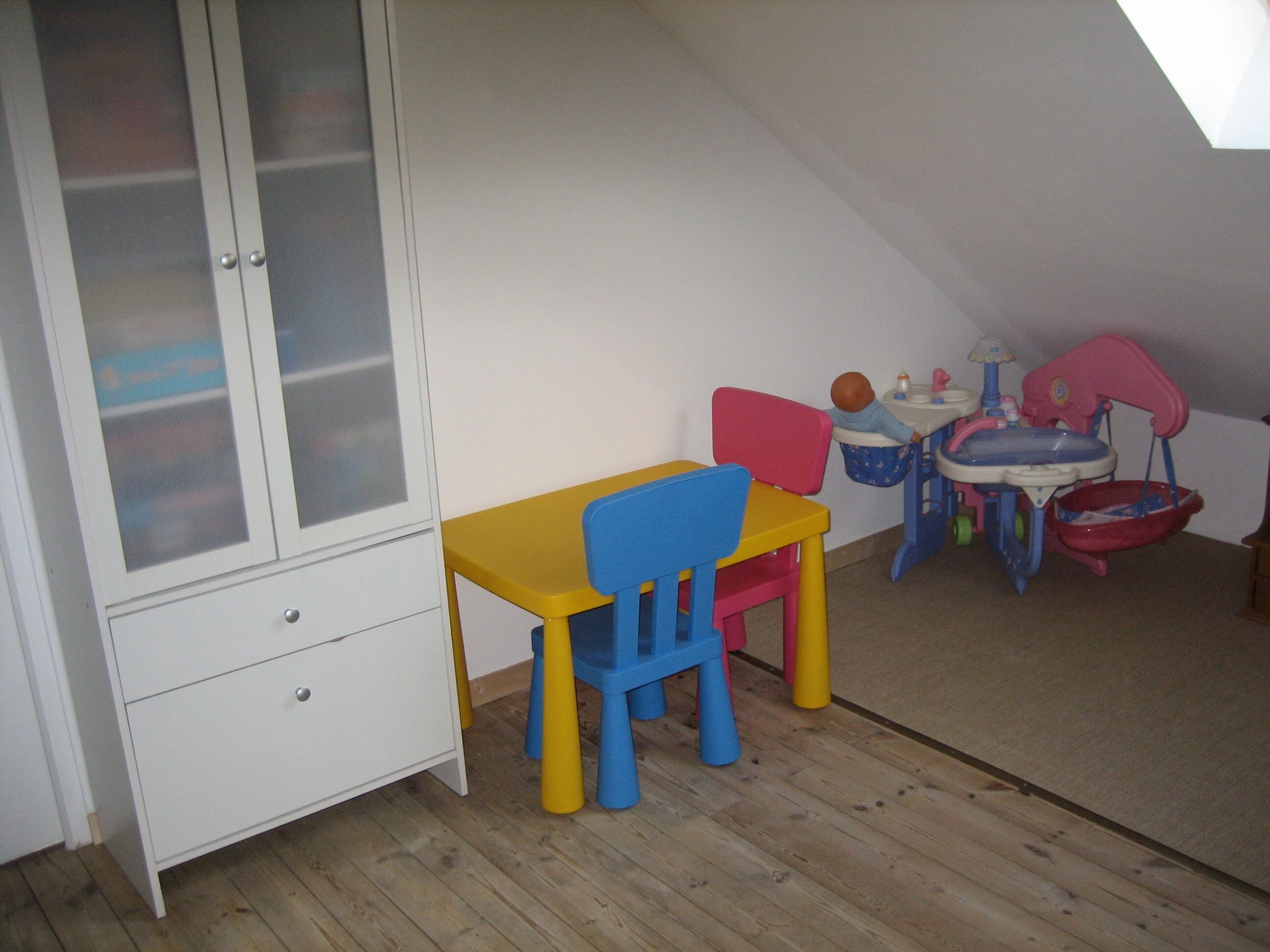 Ein Zimmer mit Holzboden, einem weißen Schrank mit Milchglasschränken, einem gelb-blauen Stuhl, einem kleinen Tisch und einem Spieltisch für Kinder mit Spüle und Spielzeug.