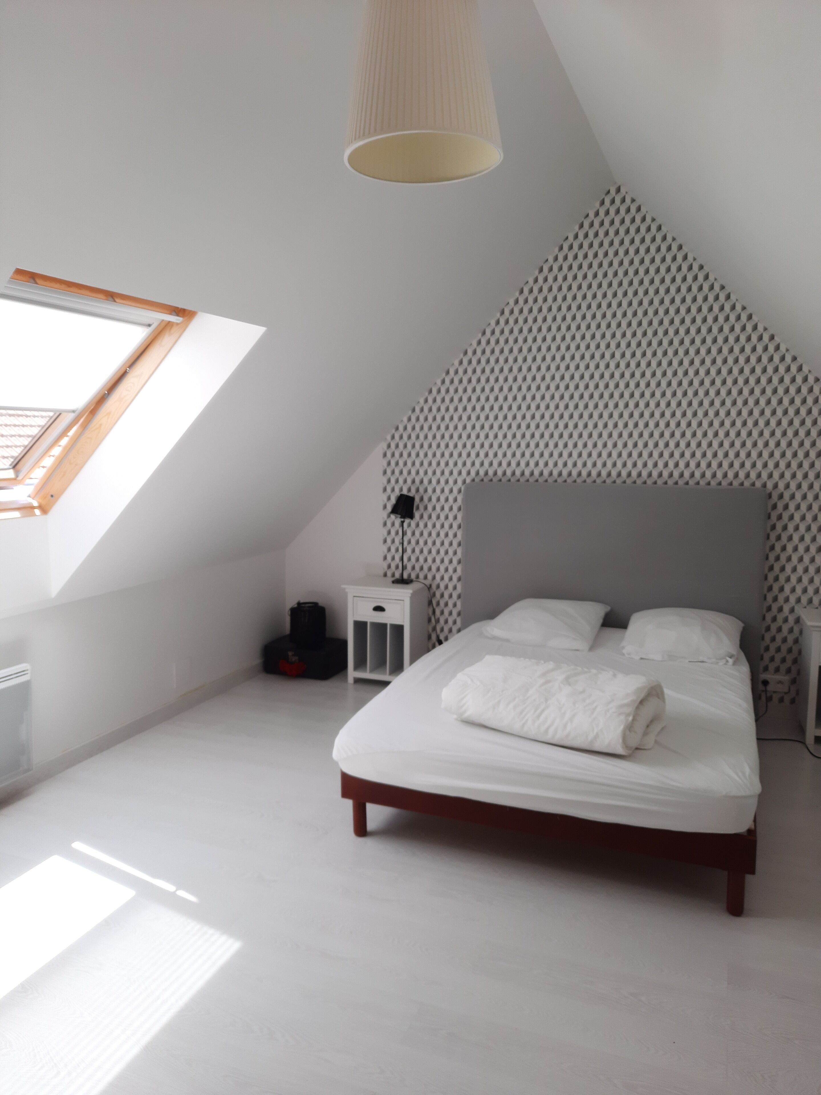 Un dormitorio con techo inclinado, una cama con ropa blanca, una mesita de noche, una lámpara y un papel tapiz estampado.