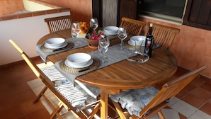 Een houten eettafel gedekt met borden, glazen en een fles, omringd door fauteuils met kussens.