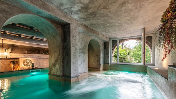 Zona d'spa amb una gran piscina interior, sostres arquejats i vista a la vegetació exterior.
