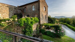 Edifici de pedra amb una torre, envoltat de vegetació exuberant i un camí de grava.