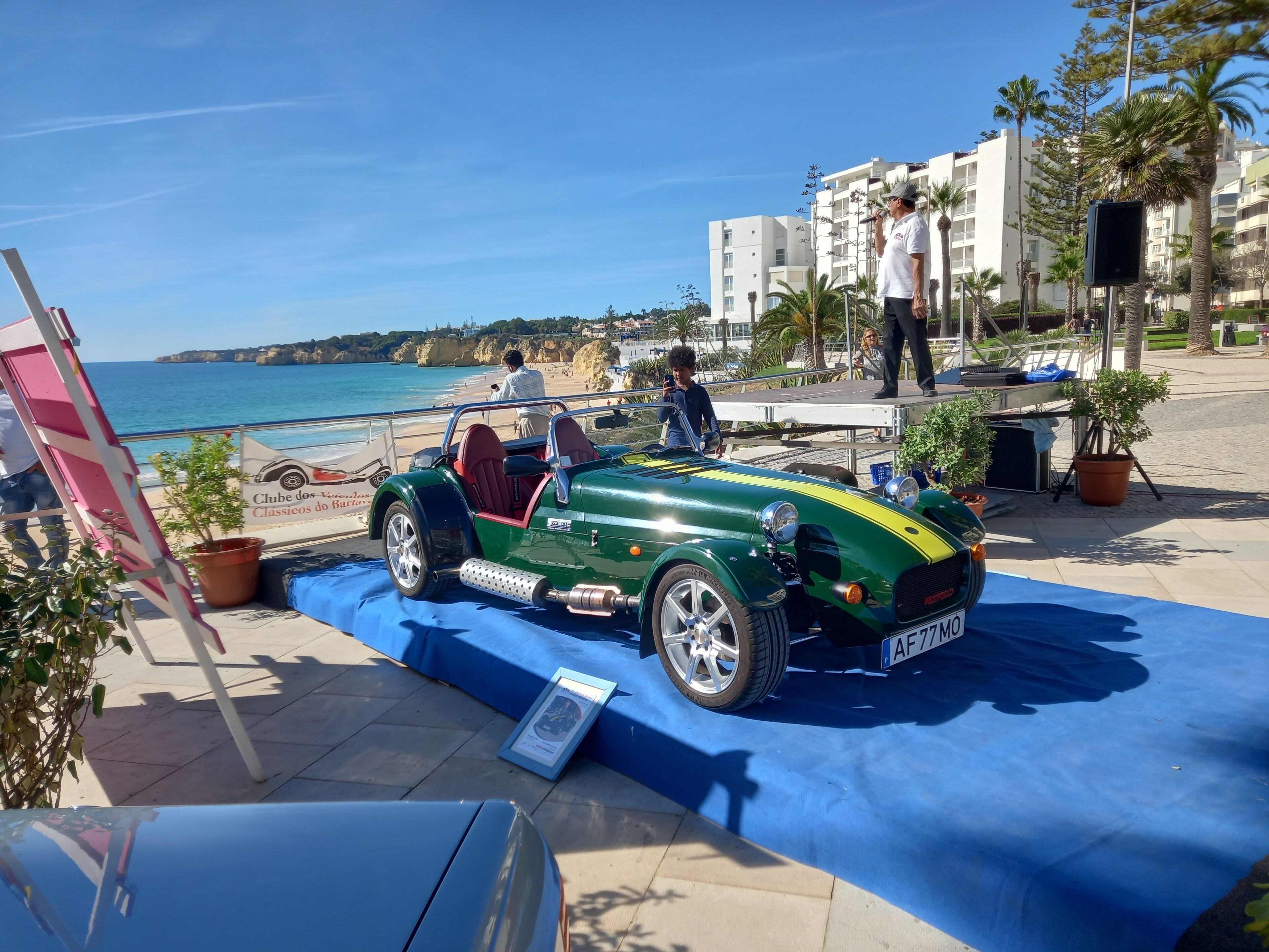 A green sports car parked on a blue carpet with a coastal backdrop.