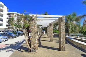 A paved walkway with stone pillars, a white pergola, and a row of parked cars.
