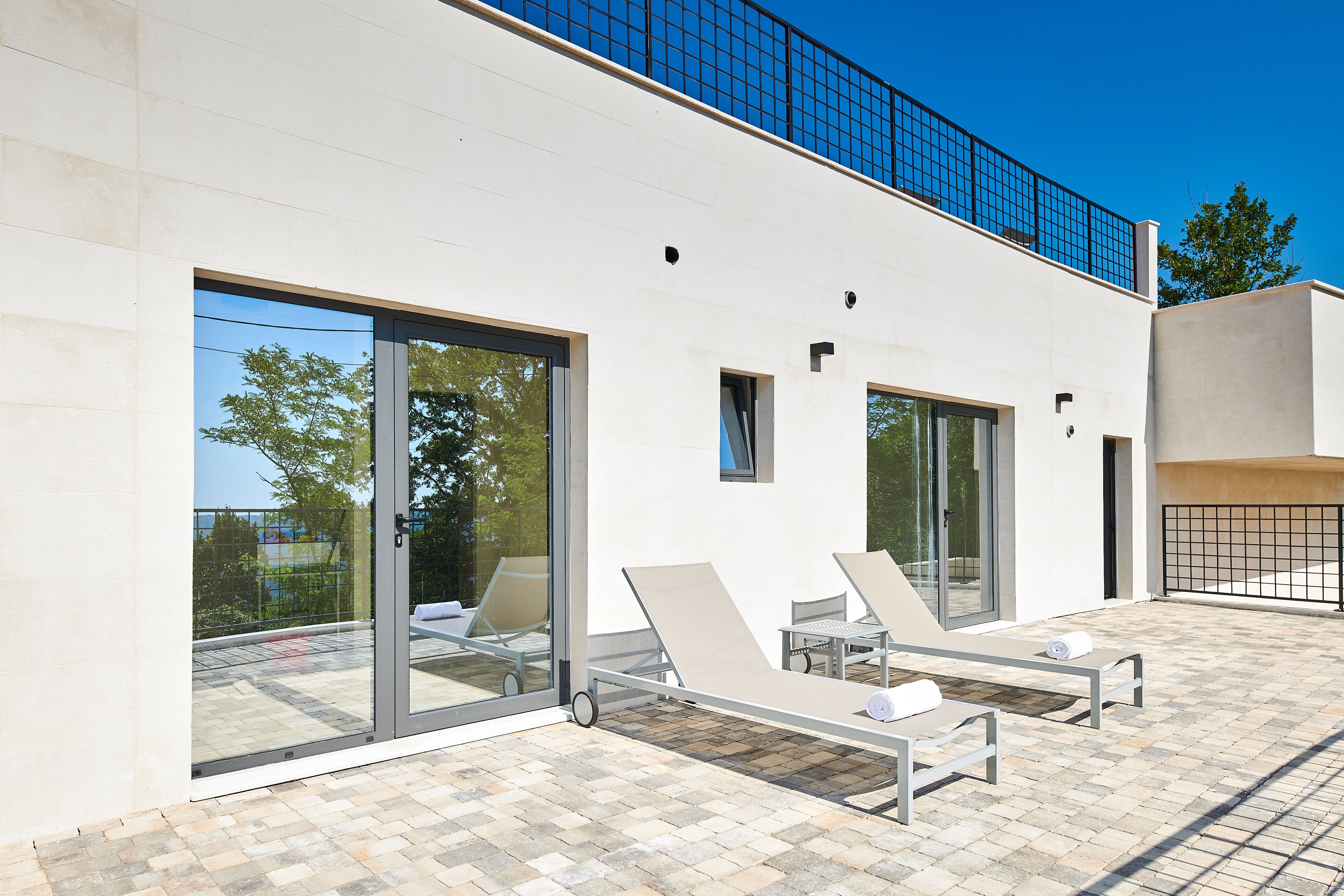 A modern outdoor lounge area with glass doors, two sun loungers, and a small table.