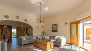 Amplia sala de estar con sofá, reposapiés y mesa de centro, un comedor con sillas y una cocina al fondo.