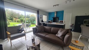 Un salon moderne avec un canapé en cuir marron, une petite table ronde et un coin repas avec une chaise. On aperçoit, au fond, une cuisine avec des armoires bleues et un comptoir noir.