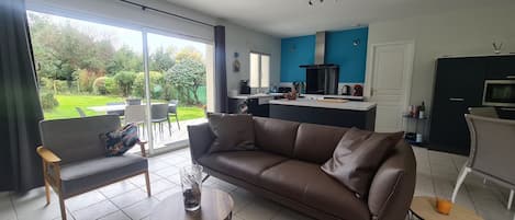 A modern living room with a brown leather sofa, a small round table, and a dining area with a chair. A kitchen with blue cabinets and a black countertop is visible in the background.