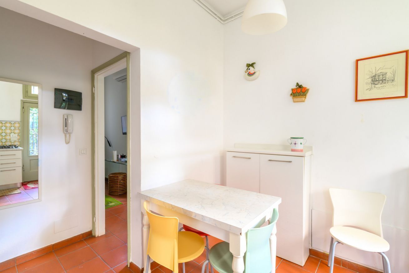 Une cuisine avec une table blanche, des chaises et un réfrigérateur. On y trouve une porte qui mène à une autre pièce, un téléphone fixé au mur et un tableau encadré.