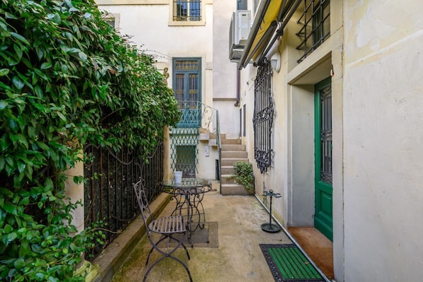 A narrow courtyard with a metal table and chair, a green door, and a staircase leading to an upper level.