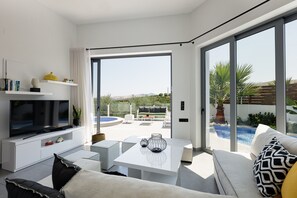 Una sala de estar moderna con televisor de pantalla plana, un sofá blanco, una mesa de centro y vistas a una piscina exterior y jardín.