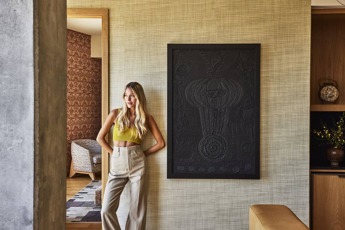A room with a large framed artwork on the wall, a wicker chair, and a wooden cabinet with a vase.