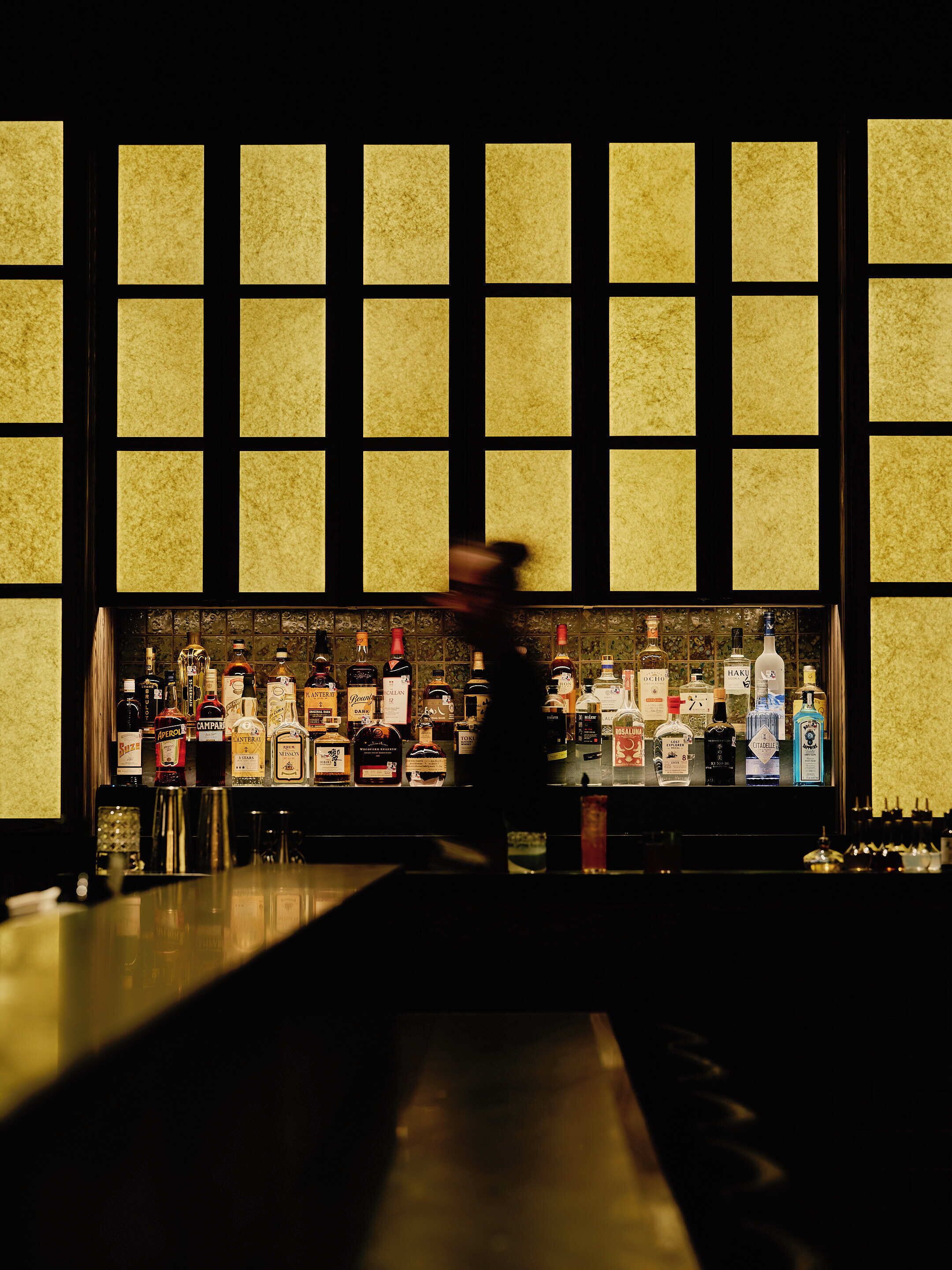 Un bar où l’on peut voir une variété de bouteilles d’alcool, et une personne qui passe devant.