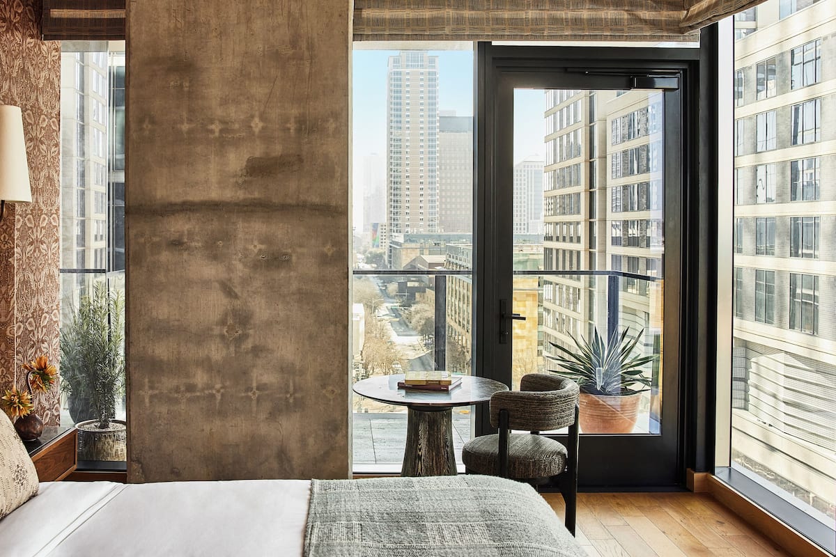 A modern hotel room with a large bed, a small round table, a chair, and a view of the city through large windows.