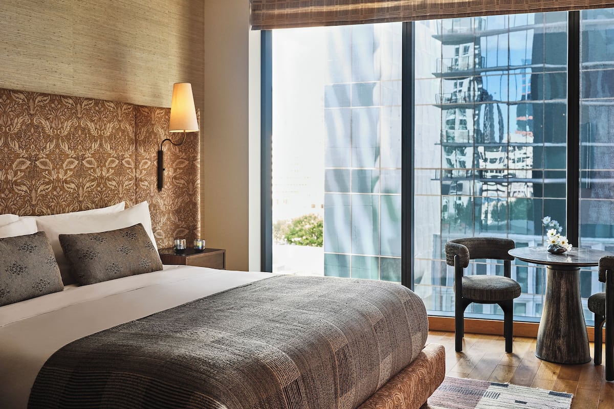 A modern hotel room with a large bed, a nightstand, a lamp, a small table with chairs, and a large window with a city view.