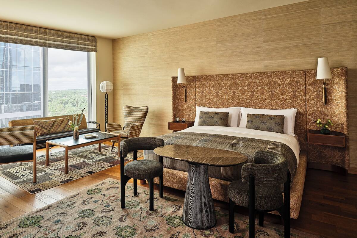 A hotel room with a large bed, a sofa, two chairs, a small table, and a view of buildings through large windows.