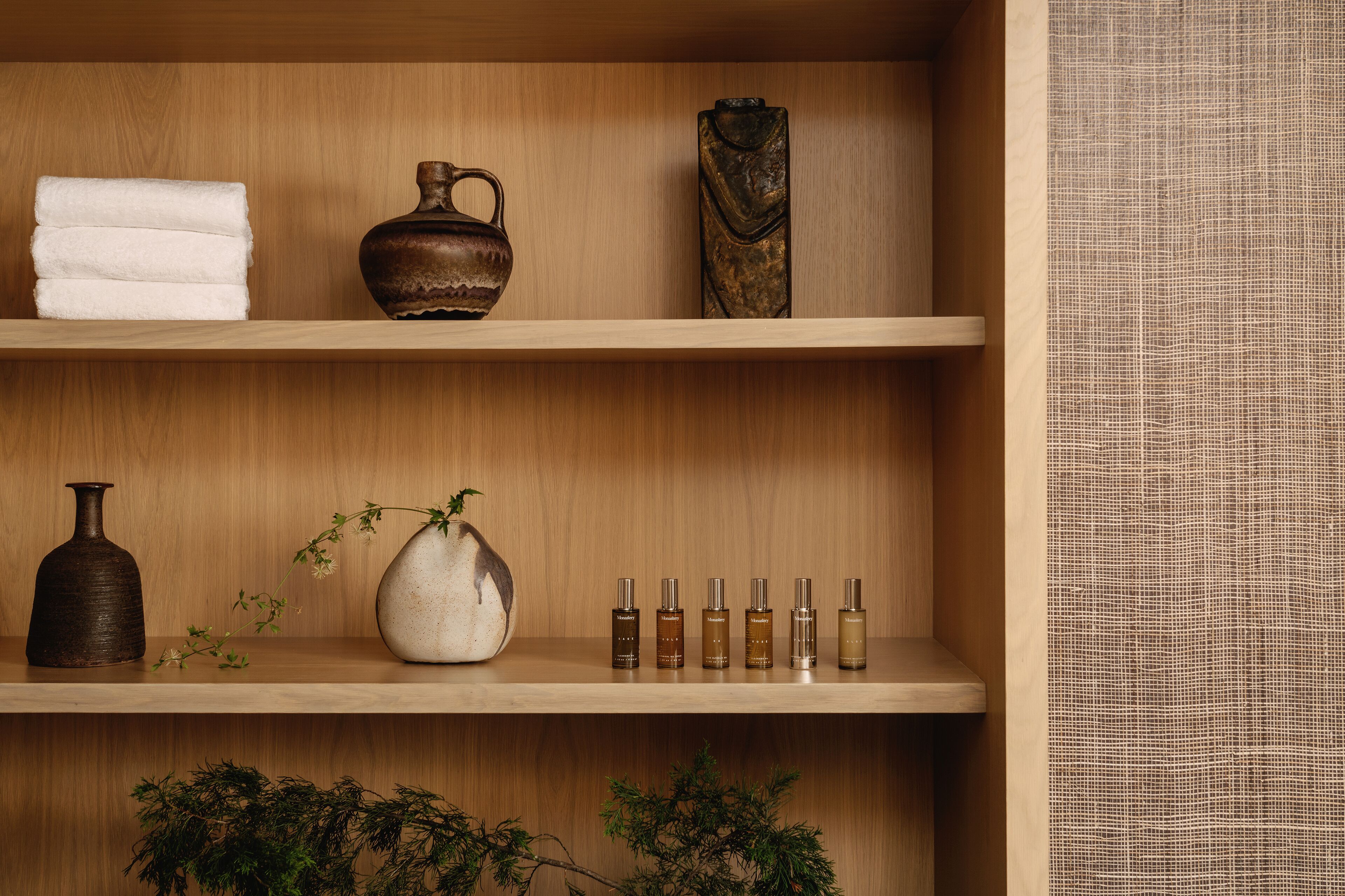 A wooden shelf with a folded towel, a vase, a decorative piece, a bottle, a potted plant, and a row of perfume bottles.