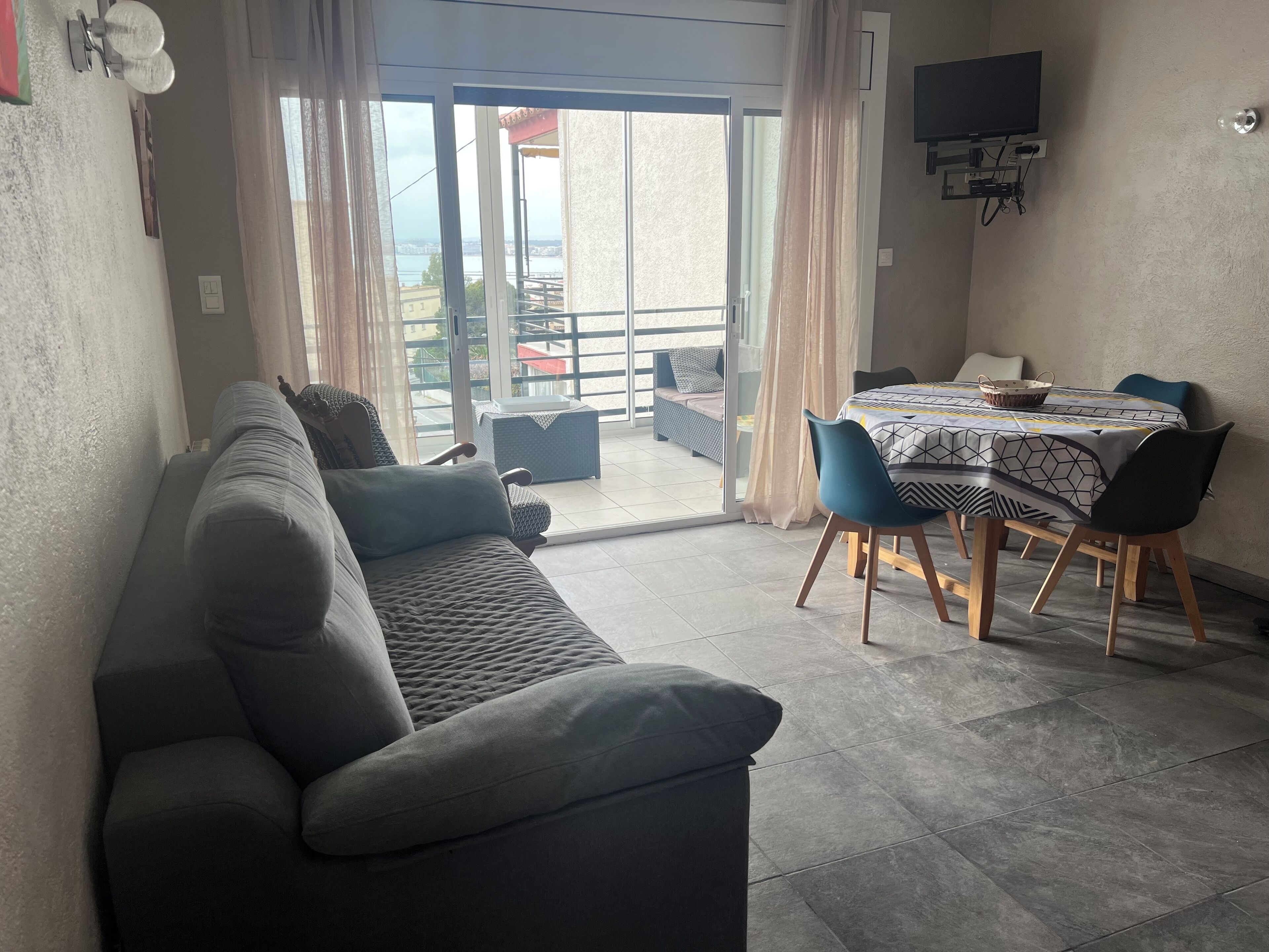 Una sala de estar con sofá, una mesa de comedor con sillas, un televisor montado en la pared y un ventanal corredero que da a un balcón exterior.