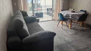 Un salon comprenant un canapé, une table à manger avec des chaises, un téléviseur fixé au mur et une porte-fenêtre coulissante donnant sur un balcon extérieur.