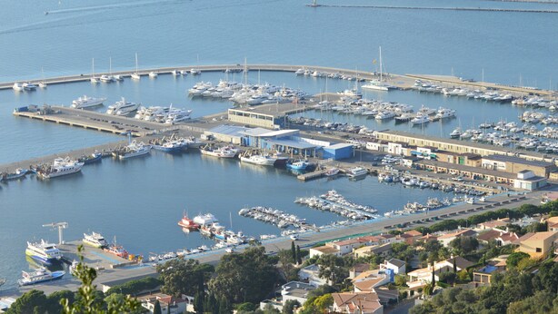 Un port de plaisance où sont amarrés de nombreux bateaux, un grand bâtiment doté d’un toit plat et une jetée qui s’avance dans l’eau.