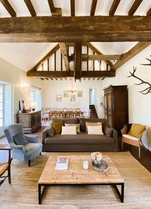 A spacious living room with exposed wooden beams, a sofa set, a coffee table, and a dining area in the background.