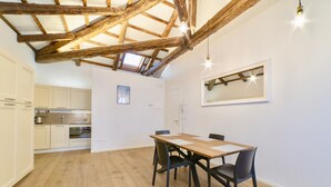 A dining area with a wooden table and chairs, a kitchen in the background, and a sloped ceiling with exposed beams. - New apartment in Murano the amazing glass island (Venezia)