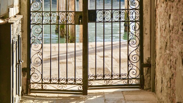 An arched metal gate with intricate designs, leading to a stone-paved pathway and a view of a canal with boats. - New apartment in Murano the amazing glass island (Venezia)