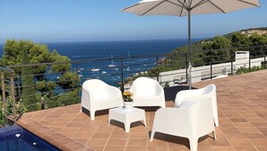 Ein Poolbereich mit weißen Stühlen und einem Tisch, einem weißen Sonnenschirm und Blick auf das Meer mit Booten.