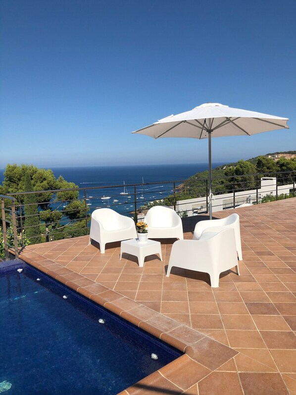 A pool area with white chairs and a table, a white umbrella, and a view of the sea with boats.