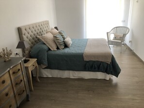Une chambre à coucher avec une tête de lit capitonnée, un lit avec une couette et un plaid de couleur turquoise et beige, une table de chevet avec des tiroirs, une lampe et un fauteuil en osier dans un coin.