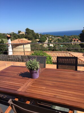 Une terrasse en bois avec une table et des chaises, un pot de fleurs et une vue sur la mer et les maisons environnantes.
