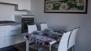 A modern kitchen with white cabinets, a gas stove, and an oven. There is a dining table with a floral tablecloth, white chairs, and a framed painting of daisies on the wall. - Apartment in residence with private garden and swimming pool for adults and children and tennis (Lazise)