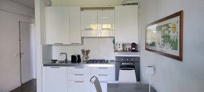 A compact kitchen with white cabinets, a gas stove, a sink, and a small dining area with a chair. - Apartment in residence with private garden and swimming pool for adults and children and tennis (Lazise)