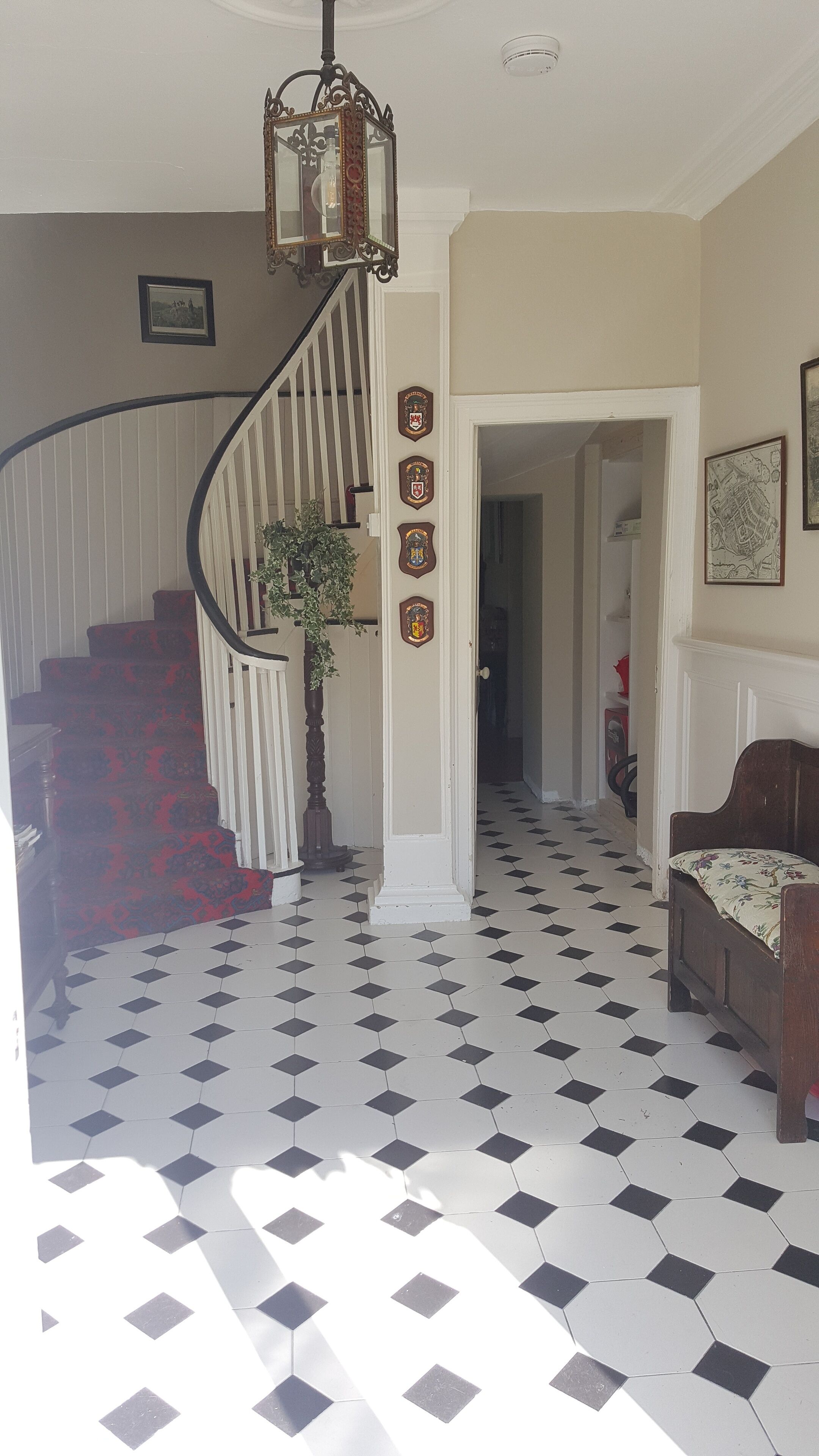 Un escalier recouvert d’une moquette rouge, avec une rampe blanche et une lanterne suspendue. Trois blasons ornent le mur, et un banc en bois, agrémenté d’un coussin à motifs floraux, est placé en contrebas.