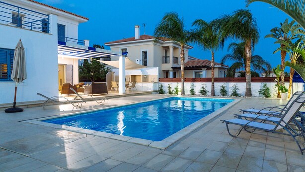 A swimming pool with lounge chairs, a covered patio area, and palm trees. - Nirvana Seaside Villa (Pervolia)