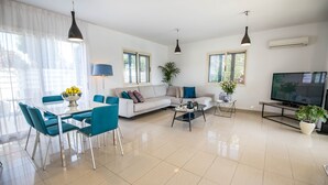 A modern living room with a sofa, dining table, chairs, and a TV. - Nirvana Seaside Villa (Pervolia)