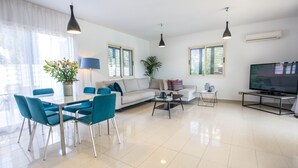 A modern living room with a sofa, dining table, chairs, a coffee table, and a TV. - Nirvana Seaside Villa (Pervolia)