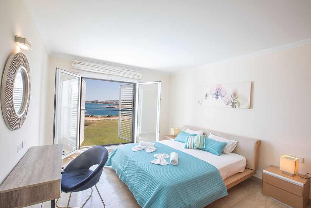 Een slaapkamer met een bed, een bureau, een stoel, een nachtkastje en uitzicht op zee.