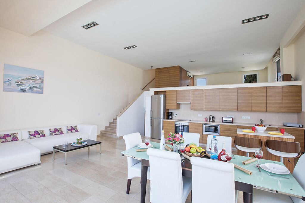 A modern living room with a dining table set for four, a white sofa, and a kitchen area with wooden cabinets and a microwave.