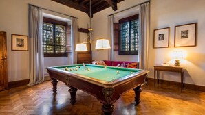 Uma mesa de bilhar em um ambiente com piso de madeira, amplas janelas com cortinas e quadros nas paredes.