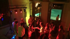 Um evento interno lotado com pessoas dançando, palco com equipamentos de iluminação e uma piscina visível ao fundo.