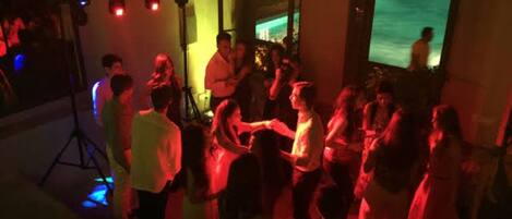A crowded indoor event with people dancing, a stage with lighting equipment, and a pool visible in the background.