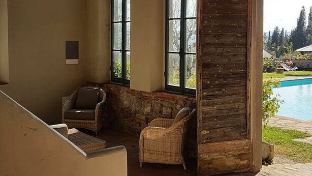 Uma área de estar coberta ao ar livre, com móveis de vime, parede de pedra e vista para uma piscina.