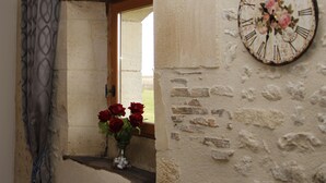 Una habitación rústica con pared de piedra, marco de ventana de madera y un reloj de pared con números romanos.