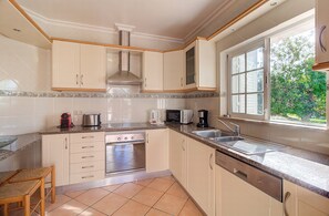 A modern kitchen with white cabinets, a stainless steel range hood, a dishwasher, and a window with a view of greenery.